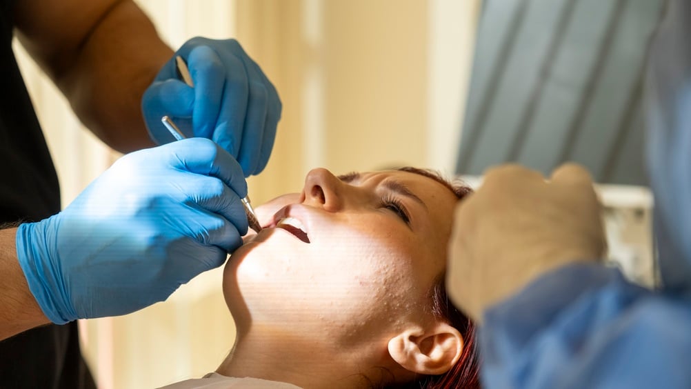 A person reclines in a dental chair with their mouth open while two gloved dentists use dental tools to examine or treat their teeth in a brightly lit room.
