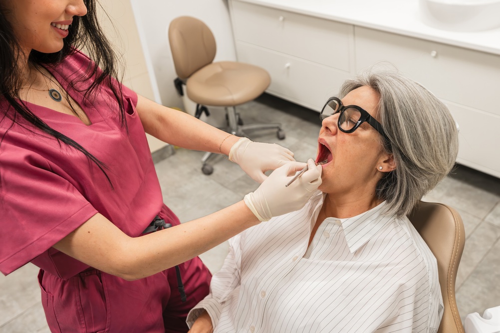 A dentist in pink scrubs examines the mouth of a seated older woman with gray hair and glasses, wearing a white shirt, in a dental office. The patient’s mouth is open, and the dentist wears gloves.