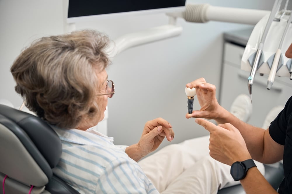 An older woman in a dental chair listens as a dentist shows her a dental implant model and explains the procedure. Dental equipment and a monitor are visible in the background.