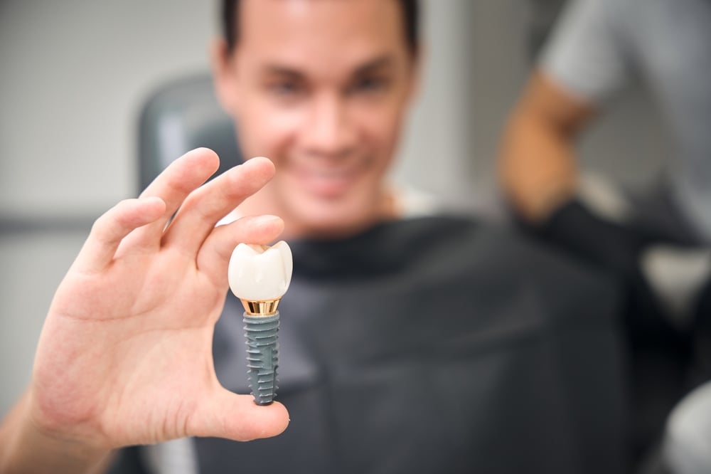 A smiling person in a dental chair holds up a large dental implant model between their fingers, showing the artificial tooth and screw. The blurred background draws attention to the implant.